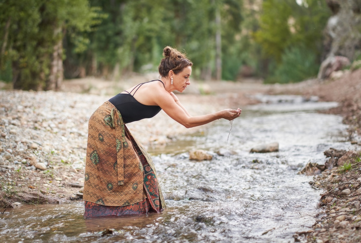 Side view full body of slim adult female in long colorful skirt standing in stream and washing raised hands against green trees in forest